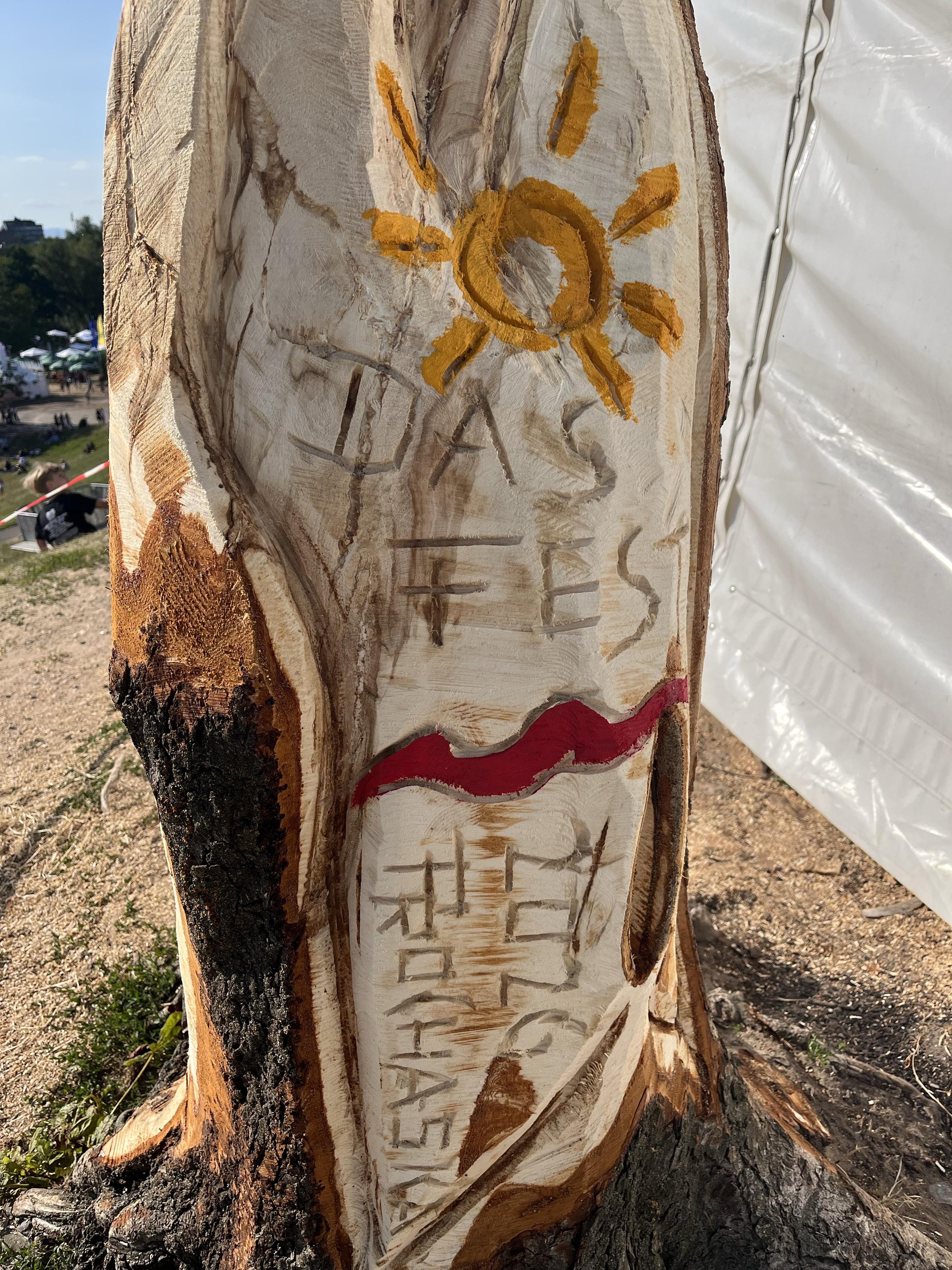 Eine Nahaufnahme eines geschnitzten Baumstumpfes mit einem gelb gestrichenen Sonnendesign und der Inschrift "DAS FEST 2026 Prochaska". Der Hintergrund zeigt eine Grasfläche mit Menschen und einem weißen Zelt.