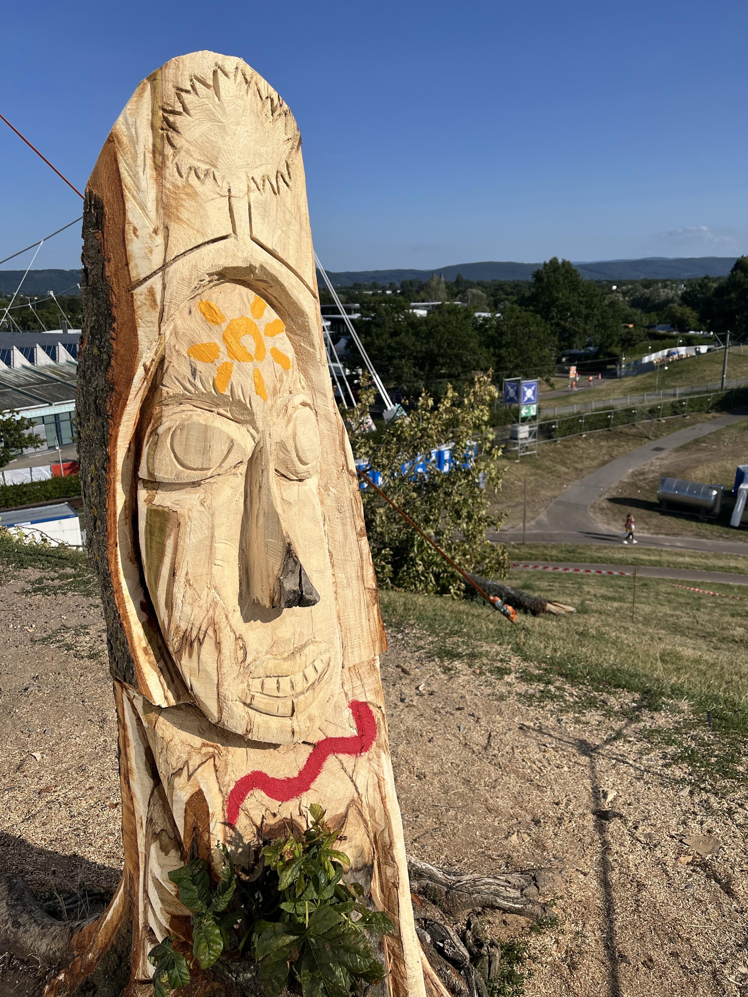 Eine geschnitzte Holzskulptur, die ein stilisiertes Gesicht mit einem Sonnensymbol auf der Stirn und einer roten welligen Linie darstellt. Aus dem Kopf scheint ein stilisierter Baum zu wachsen, was den Eindruck erweckt, dass es das Gesicht des Mount Klotz ist.
Die Skulptur befindet sich auf einem grasbewachsenen Hügel mit einem klaren blauen Himmel und fernen Bäumen im Hintergrund.